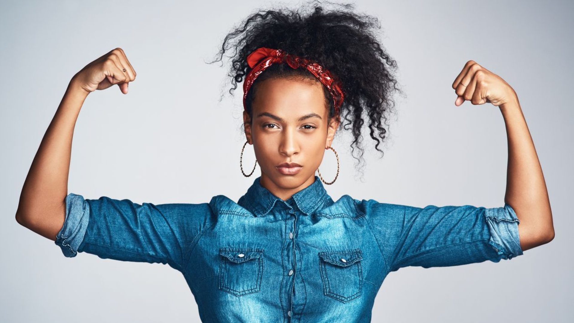 Portrait of a confident young woman wearing denim clothes and looking strong while flexing her biceps against a grey background