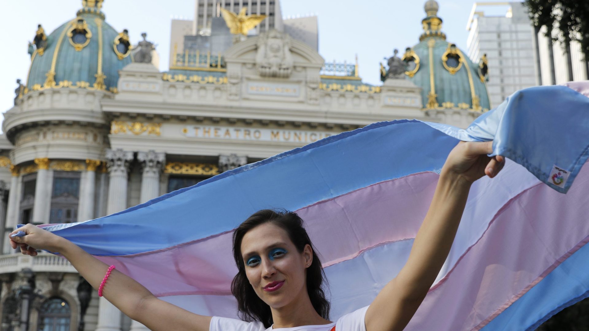 A cantora lírica e pianista Vivian Fróes, mulher trans, posa com  bandeira em frente ao Theatro Municipal, na Cinelândia.